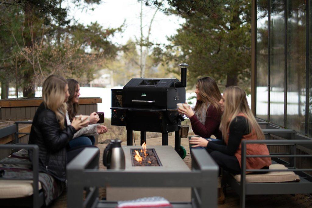 four women sitting on an outdoor patio around a table with a gas fireplace insert and a smoker BBQ with wood pellet attachment in the background